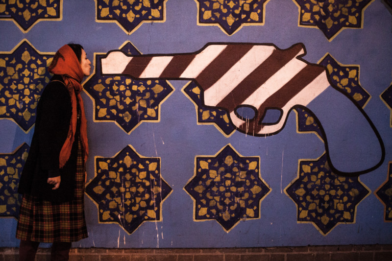 Symbolic portrait of young woman giving a kiss to a gun painted in the colors of USA on old closed US embassy in Tehran.
