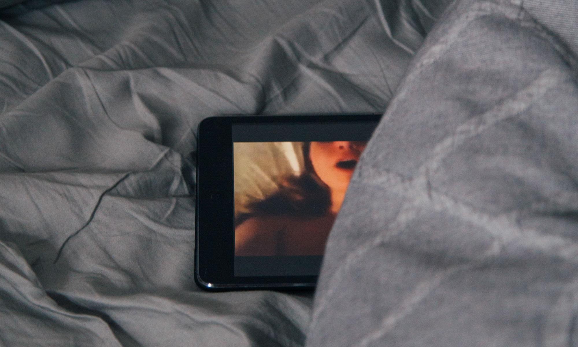 Smartphone liegt auf dem Bett, auf ihm ist eine stöhnende Frau zu erkennen. Das Handy wir teilweise von der grauen Bettwäsche bedeckt.