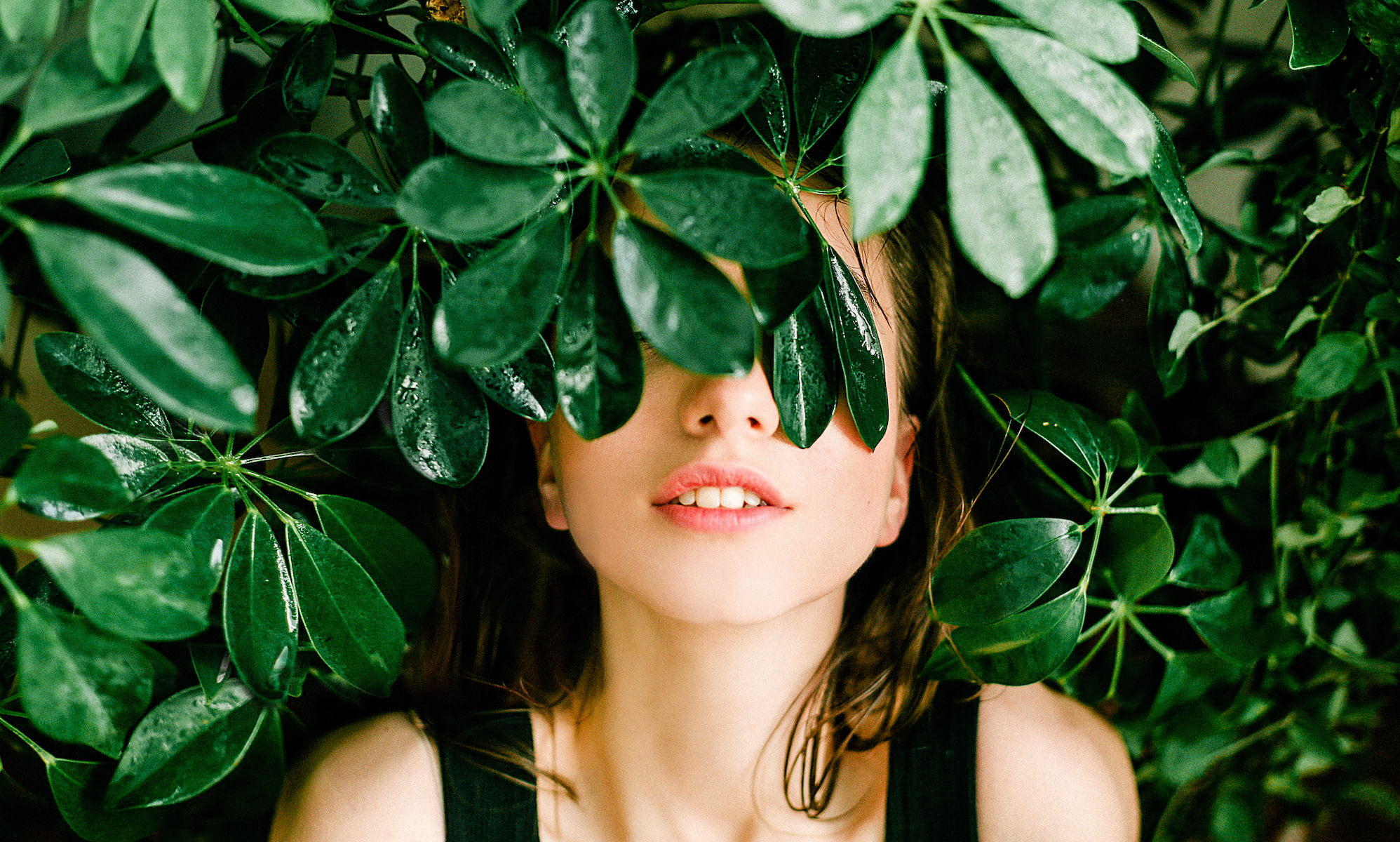 Junge Frau liegt mit dem Gesicht hinter einem Blatt. Im Hintergrund sind Pflanzen.