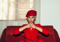 Frau in rotem Blazer sitzt auf einer roten Couch und schaut selbstbewusst in die Kamera