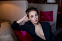 Salomé Balthus lehnt in schwarzem Oberteil mit zurückgegelten Haaren an einem Kissen mit einem Sofa im Hintergrund