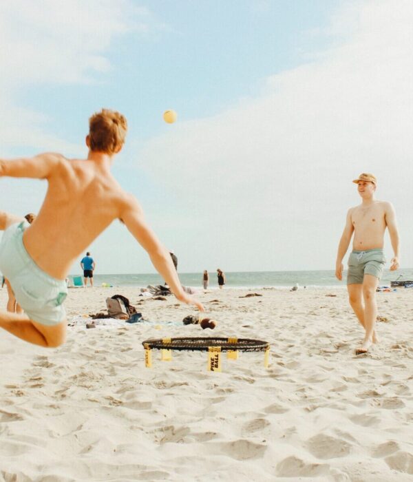 Ein paar Menschen spielen Roundnet am Strand mit einem Trampolin in der Mitte, einer davon springt mit dem Rücken zur Kamera nach hinten