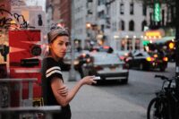 Frau lehnt an einem Gebäude, hat ein Handy in der Handy. Schaut genervt in die Kamera.