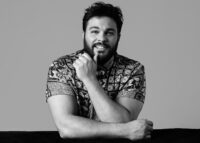 Ein junger Mann mit dunklen Haaren und Bart lächelt in die Kamera. Er stützt sich auf seinem Arm auf. Das Foto ist schwarz-weiß.