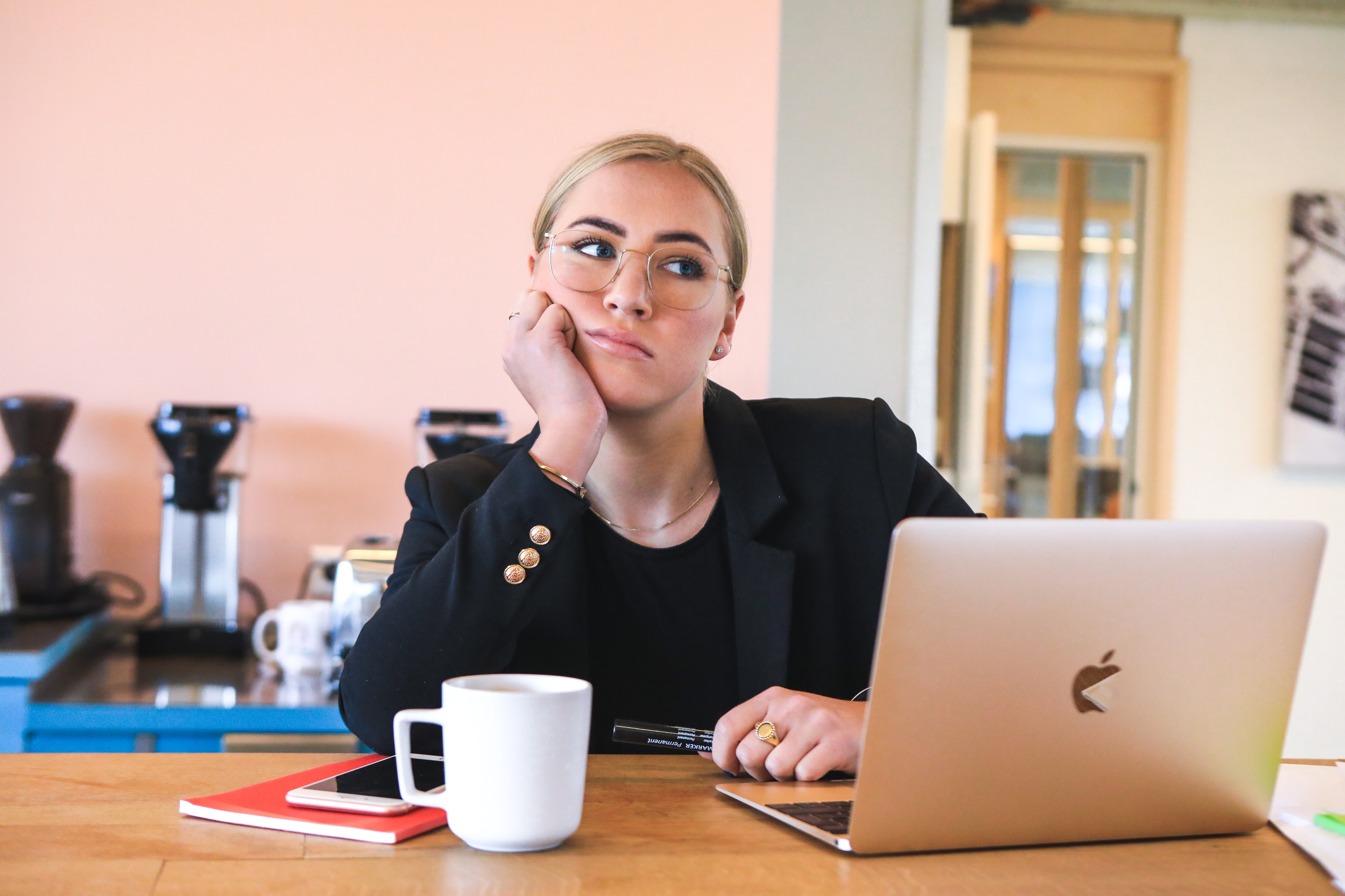 Eine junge Frau mit blonden Haaren und Brille sitzt vor einem Laptop und schaut nachdenklich und etwas missmutig ins Leere