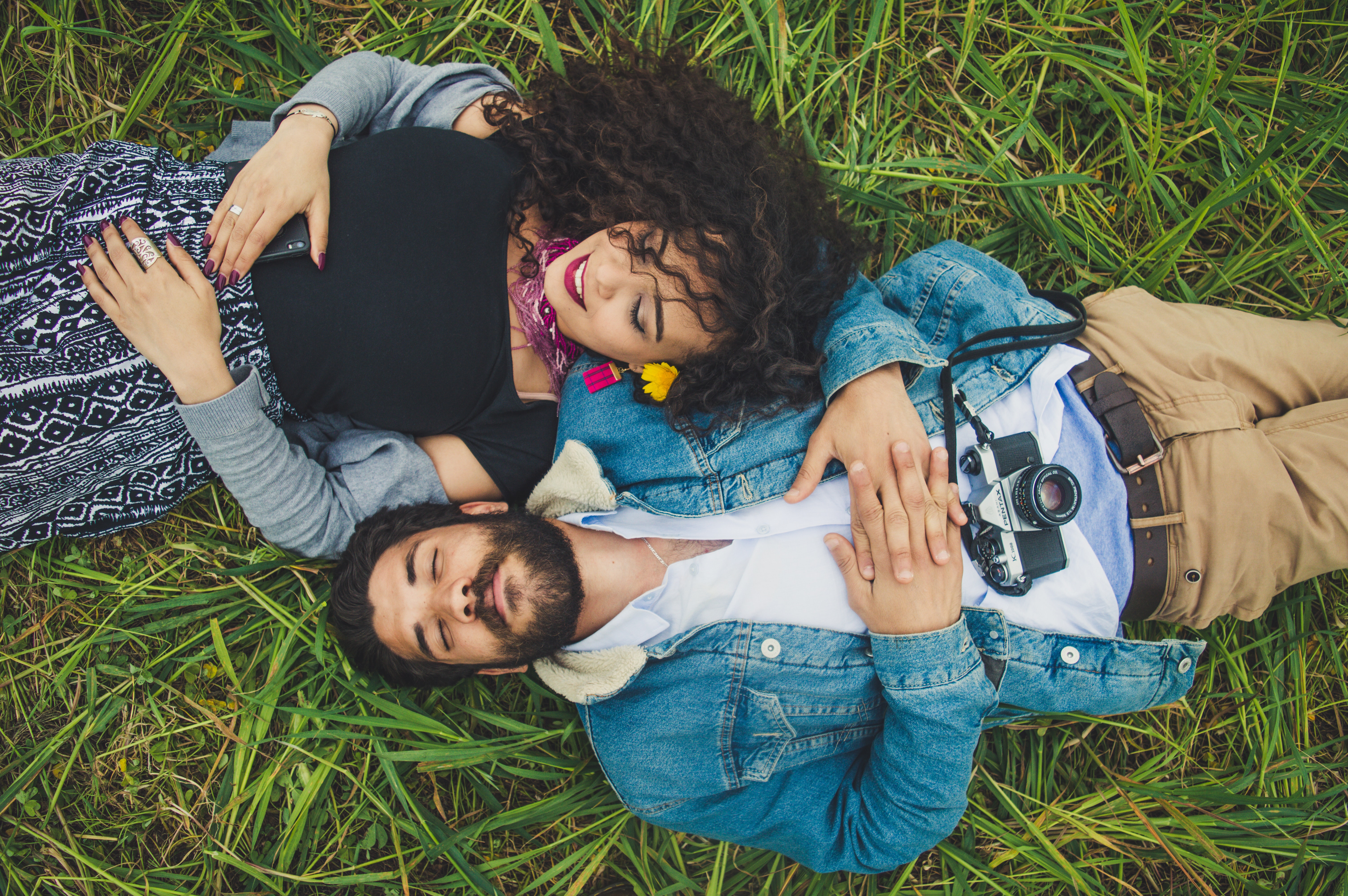 Eine Frau mit lockigen, braunen Haaren und ein Mann mit braunen Haaren und Bart liegen lächelnd im Gras. Ihre Köpfe liegen nebeneinander, die Füße zeigen in unterschiedliche Richtungen.