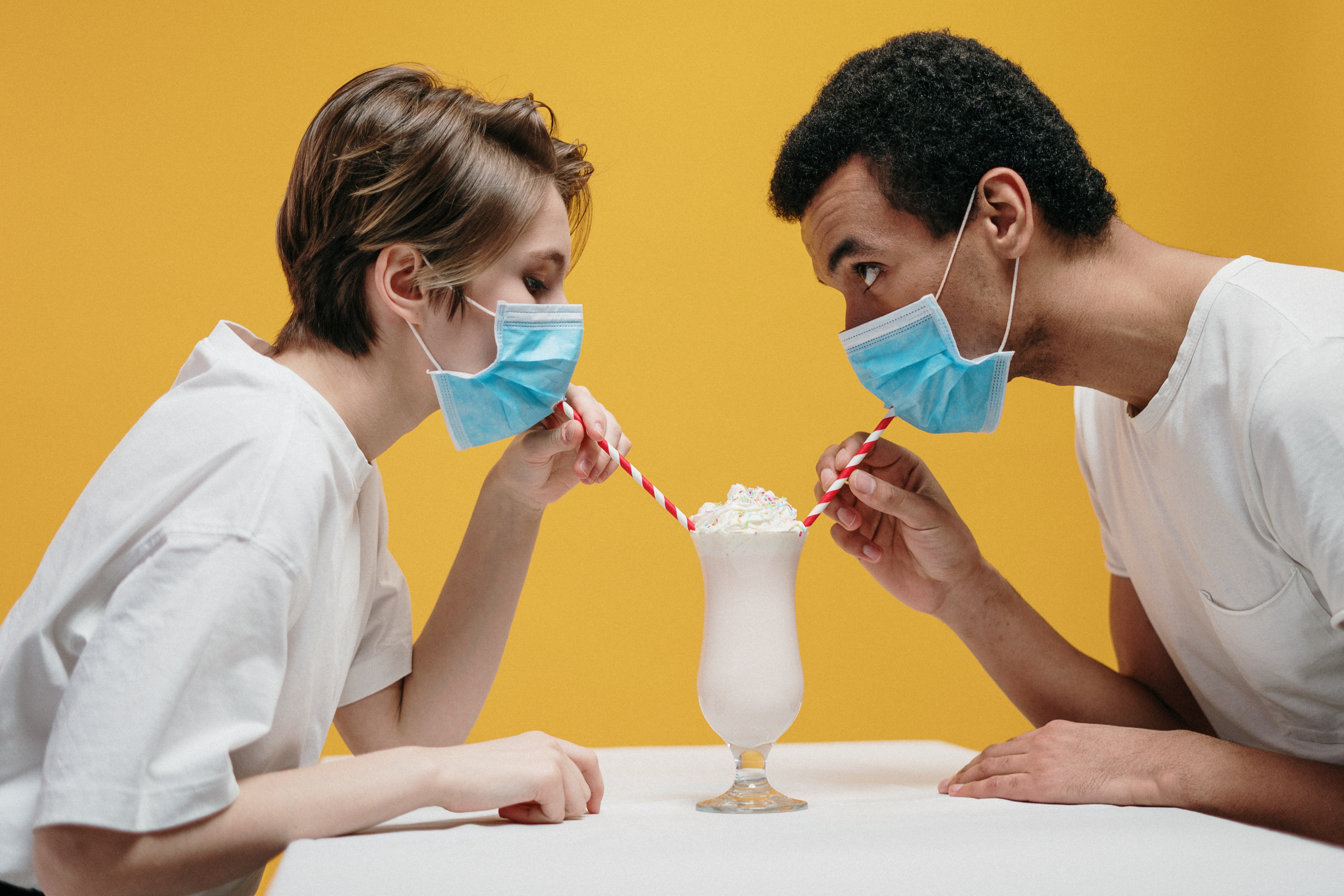 Eine Frau und ein Mann sitzen sich an einem Tisch vor einem gelben Hintergrund gegenüber. Sie tragen einen Mund-Nasen-Schutz und trinken gemeinsam mit Strohhalmen einen Milchshake.