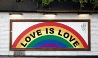 Man sieht ein Schild an einer weißen Backsteinwand, auf dem ein Regenbogen abgebildet ist. Auf dem Regenbogen steht in schwarzer Schrift "Love is Love"