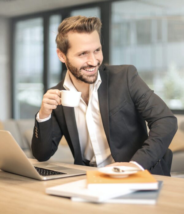 Ein Mann im Anzug sitzt an einem Tisch, trinkt Kaffee und lächelt.