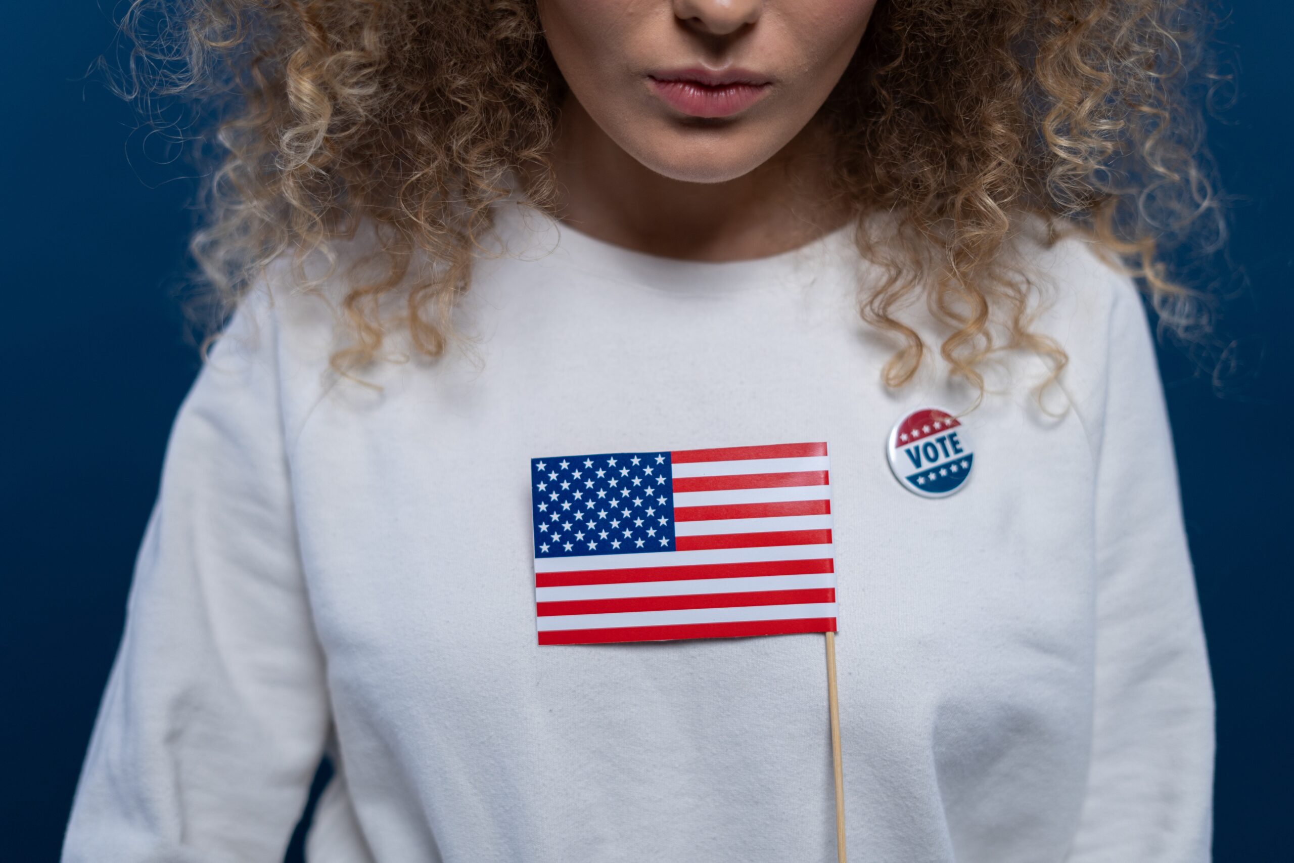 Eine junge Frau mit weißem Pulli, auf dem ein "I voted"-Sticker klebt, hält eine kleine US-amerikanische Fahne in der Hand