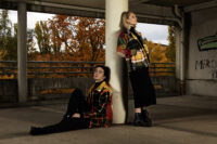 Ein Mann und eine Frau lehnen an einer Säule. Sie tragen bunte Jacken, Mode des Labels MOOT