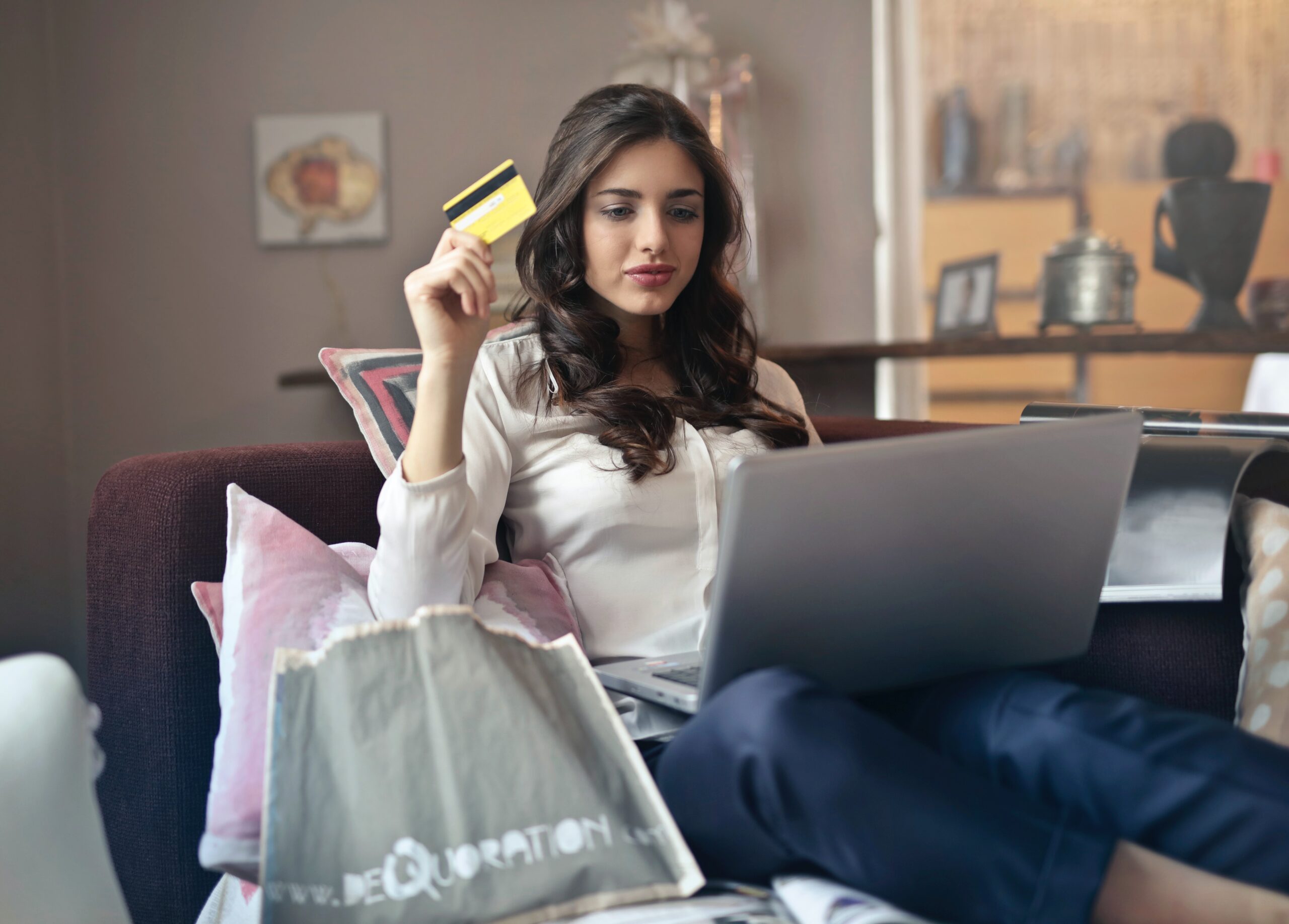 Eine Frau sitzt auf der Couch vor einem Laptop. Sie hält eine Kreditkarte in der Hand.