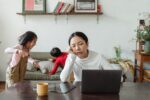 Eine Frau sitzt müde vor einem Laptop, im Hintergrund spielen Kinder