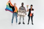 Drei Menschen mit Regenbogenflaggen und Demo-Plakaten