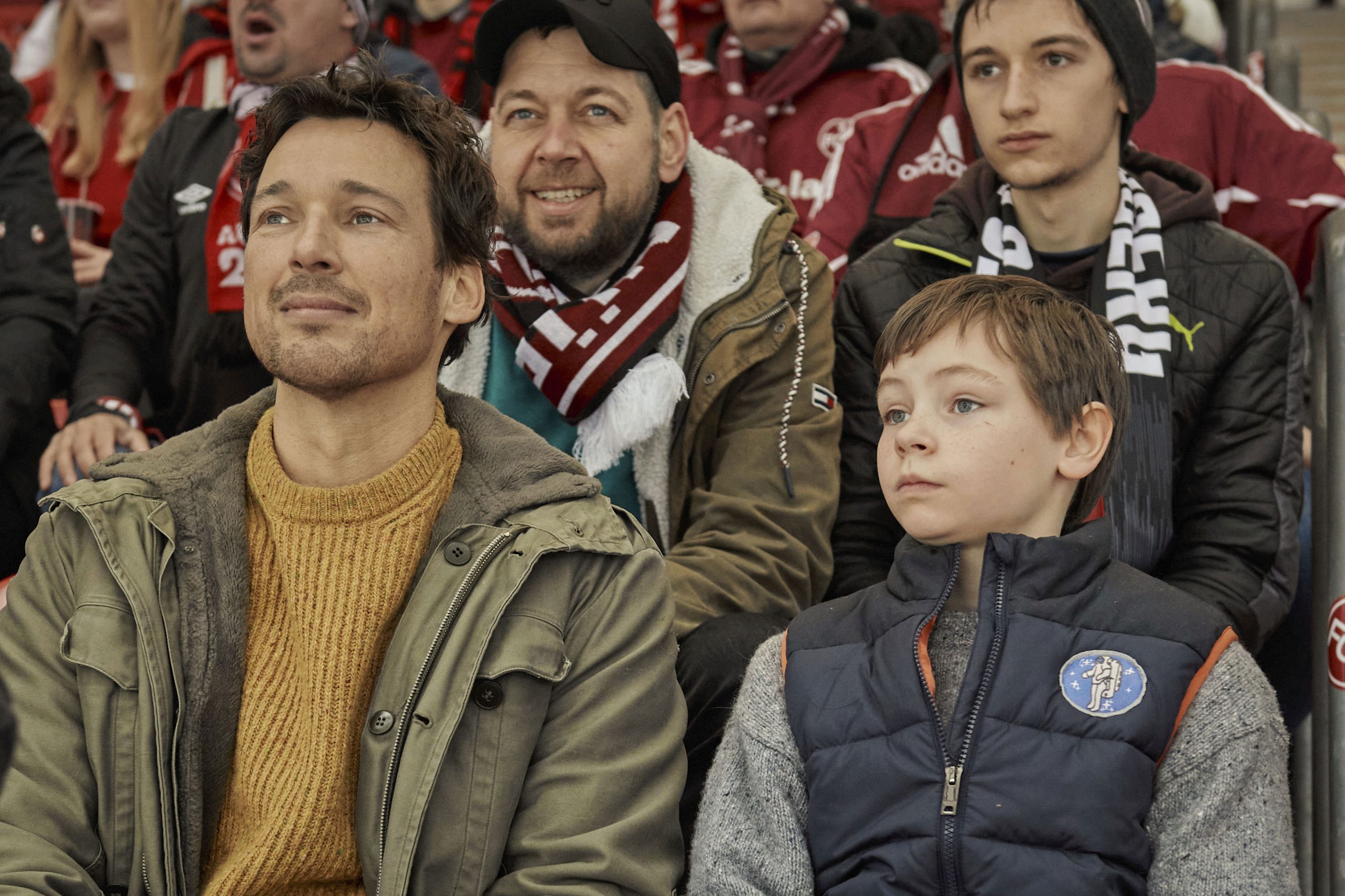 Florian David Fitz als Mirco mit seinem Sohn Jason (gespielt von Cecilio Andresen) im Stadion. Dahinter sitzen der "echte" Mirco und Jason, auf deren Geschichte der Film beruht. © Leonine Studios